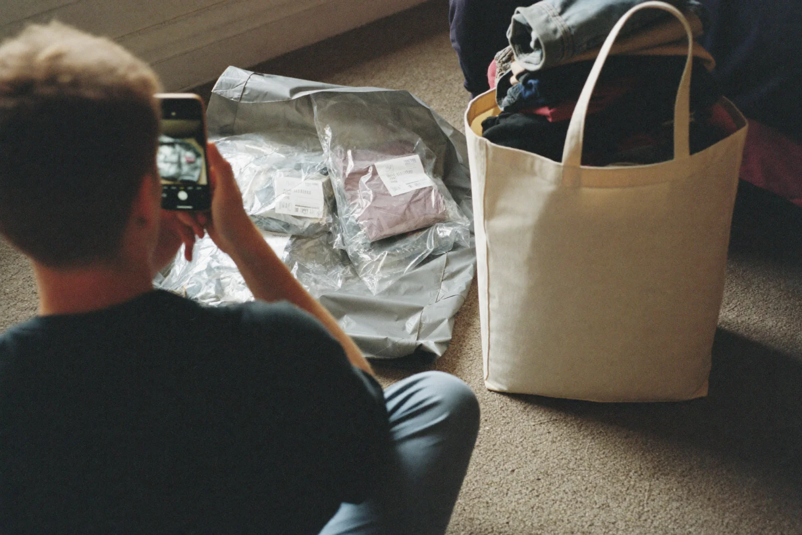 A young adult sitting on a bedroom carpet, phone raised to film an opened grey mailer spilling new clothes still sealed in labelled polybags, with a plain canvas tote of folded clothes standing beside it