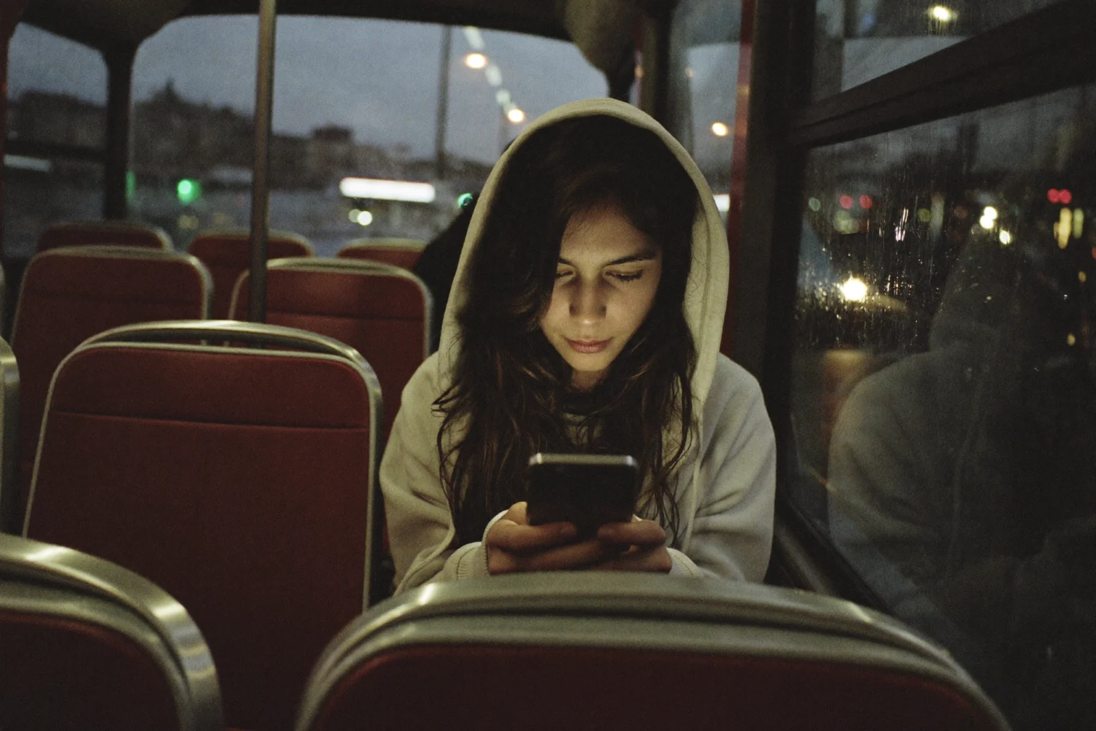 A young adult in a hoodie sitting alone among empty seats on the upper deck of a red double-decker bus at dusk, face lit from below by their phone screen, rain-streaked window and blurred city lights behind them