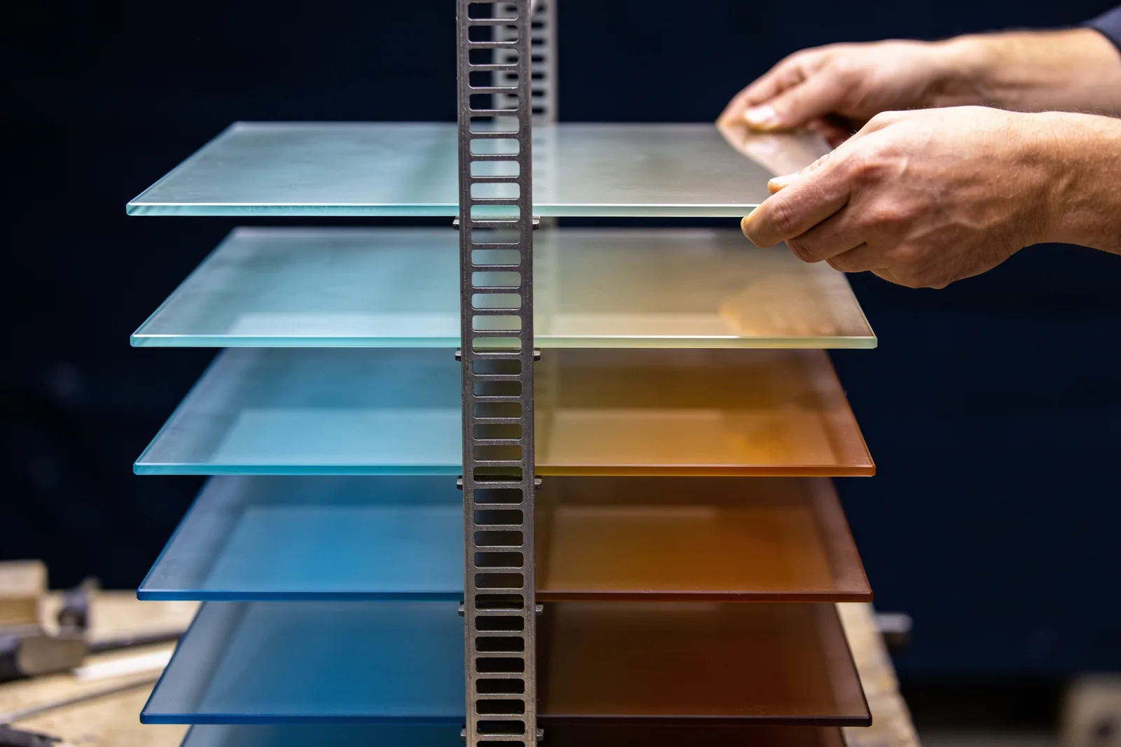 Hands lowering translucent glass panels tinted blue and amber into a slotted steel frame, stacking into one tower