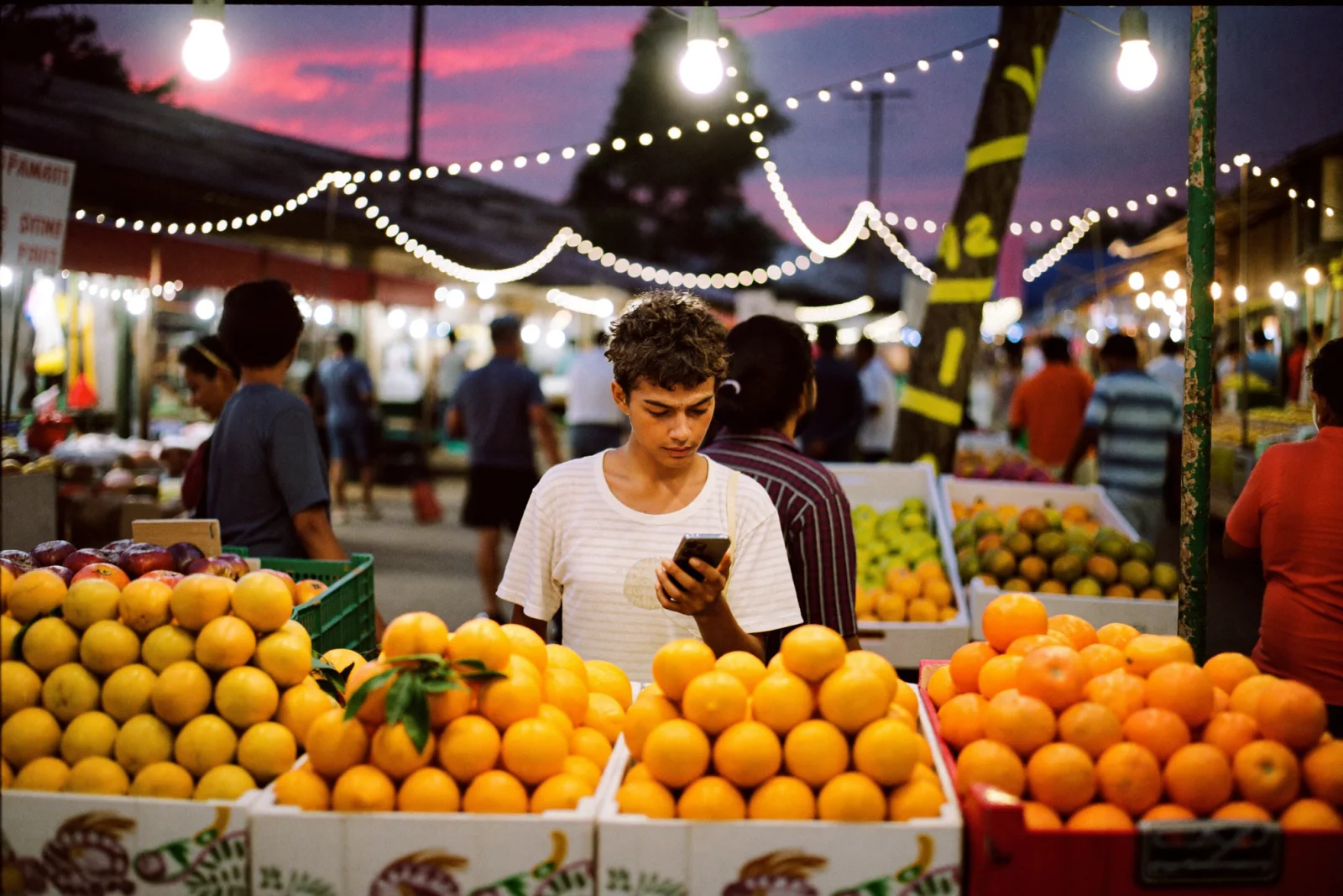 A vendor checking a phone at a busy evening street market lit by strings of bulbs, stalls stacked with oranges, illustrating how central Instagram is to daily life in emerging markets